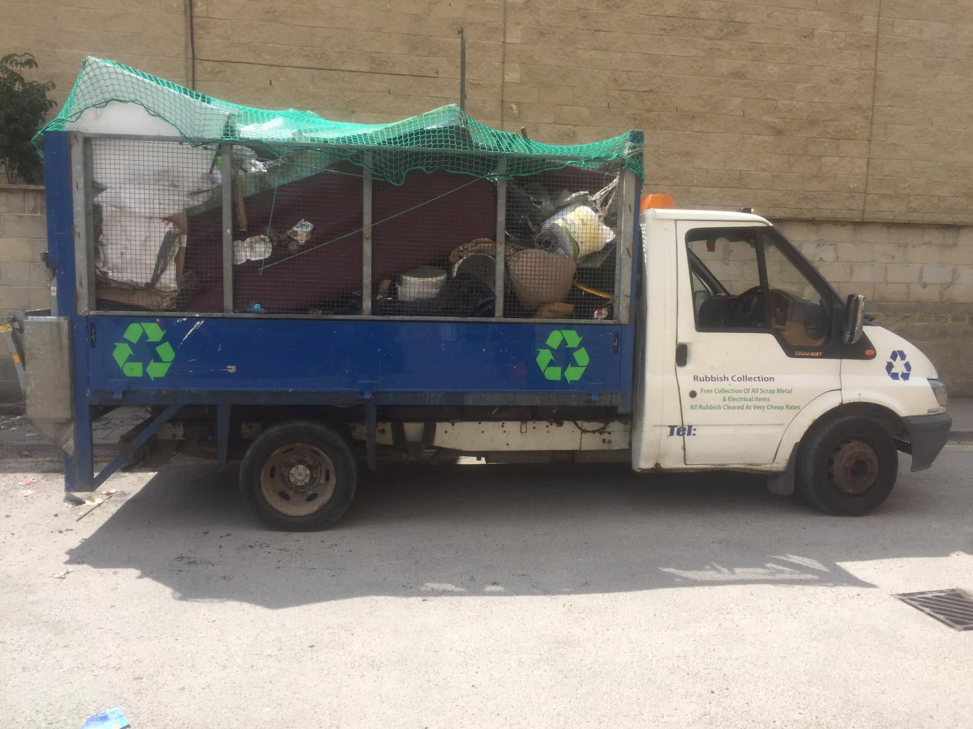 Blue and white rubbish collection truck loaded with bulky waste beside a stone wall